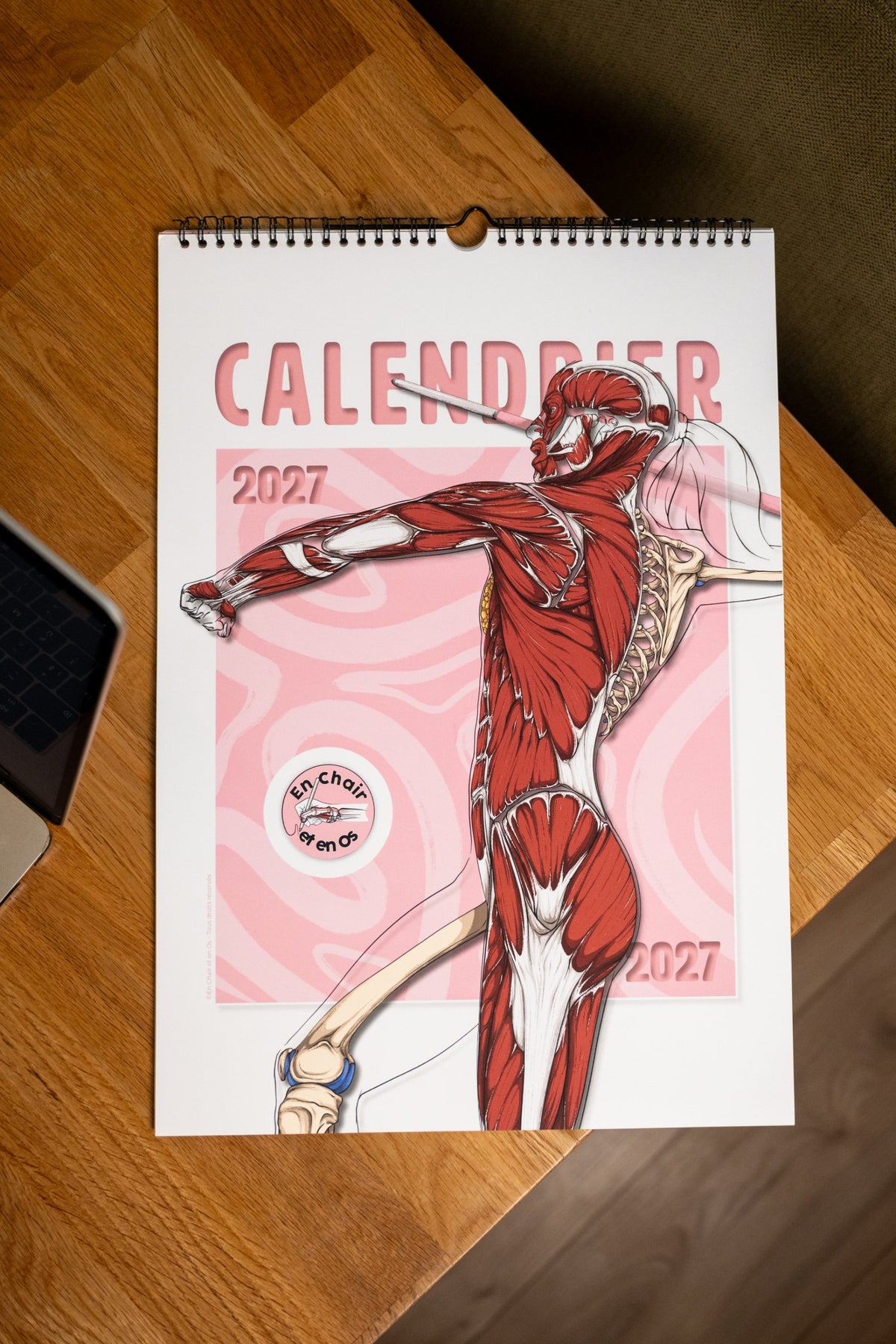 Calendrier mural A3 2027 En Chair et en Os posé sur une table en bois, couverture avec l'écorché de la lanceuse de javelot